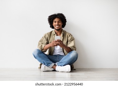 Online Communication. Happy Black Man Messaging On Smartphone While Relaxing On Floor Near White Wall Indoors, Young African American Man Looking At Mobile Phone Screen And Smiling, Copy Space