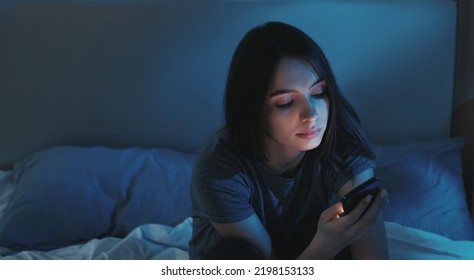 Online Communication. Sleepless Night. Pretty Smiling Woman Sitting O Bed Scrolling Smartphone In Light Shadow Room Interior.