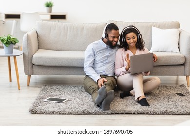 Online Communication. Smiling African American Man And Woman Making Digital Video Chat With Friends Or Family Using Laptop, Waving To Webcam, Wearing Headset, Sitting On The Floor Carpet At Home