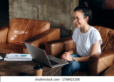 Online Communication. Young Attractive Girl With A Glasses Sits In An Armchair Indoors And Communicates With Clients Via Video Conference Discussing Working Moments Or Learning Online