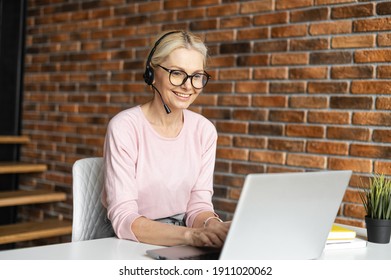 Online Consultant In Touch. A Middle-aged Woman Wearing Headset Talking On The Distance With A Customers, Clients Sitting At The Desk With A Laptop In A Modern Office Space