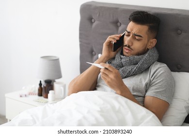 Online Consultation. Sick Arab Man Wrapped In Scarf Holding Thermometer And Calling Doctor On Phone While Sitting In Bed At Home, Ill Middle Eastern Man Suffering Cold Or Fever, Free Space