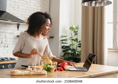 Online cooking tutorial. African young woman teenage girl using laptop for vlogging blogging looking for recipe while cooking vegetable vegan salad meal, healthy eating habits. - Shutterstock ID 2108600048