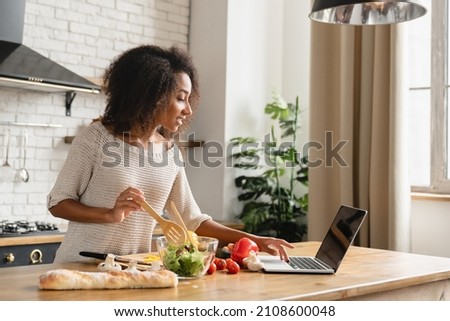 Online cooking tutorial. African young woman teenage girl using laptop for vlogging blogging looking for recipe while cooking vegetable vegan salad meal, healthy eating habits.