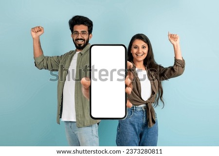 Online Coupons, Promo Codes, Cash Back. Emotional excited young indian couple showing big phone with white blank screen and clenching fists, isolated on blue background, mockup, copy space