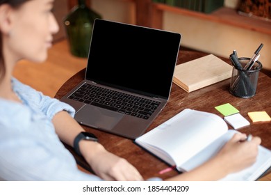 Online Course Concept. Over The Shoulder View Of Female Student Sitting At Table And Writing In Notebook, Using Laptop With Black Empty Screen For Mock Up Template, Watching Lecture Or Webinar