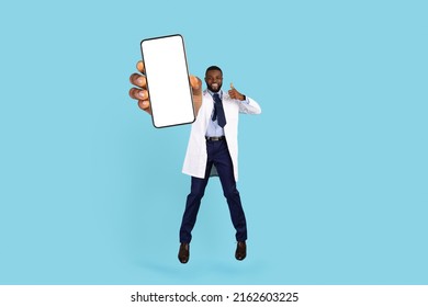 Online Doctor. Black Therapist In Uniform Jumping With Big Blank Smartphone In Hand And Showing Thumb Up At Camera, Smiling African American Physician Recommending New Mobile App, Mockup