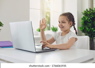 Online Education Or Communication. Waving Arms Child Girl Gretting Looks In Laptop Sitting At Table In Room