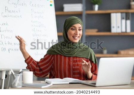 Online Education Concept. Portrait of smiling female arabic teacher in hijab pointing at whiteboard, explaining English grammar rules to students. Excited woman looking at laptop screen, talking
