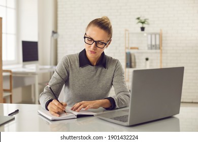 Online Education Concept. Serious Female Student Studying, Watching Webinar, Taking Notes In Notebook. Woman In Glasses Sitting At Desk With Laptop At Home Or In Office And Writing In Business Planner