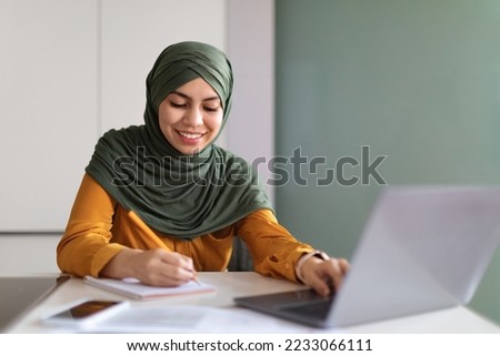 Online Education Concept. Young Smiling Muslim Woman In Hijab Study With Laptop At Home, Islamic Lady Using Computer And Taking Notes To Notepad, Enjoying Distant Learning, Free Space