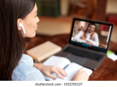 Online education, e-learning concept. Young woman with earbuds having video conference with male teacher, sitting at desk and using laptop, taking notes, discussing studying material, home interior