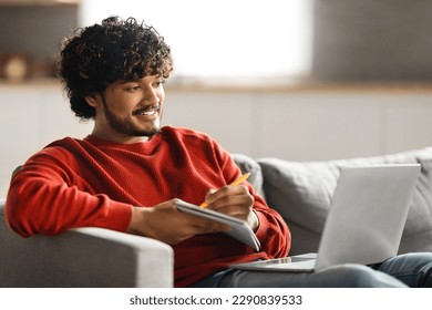 Online Education. Handsome young indian man using laptop and taking notes while sitting on couch at home, smiling eastern guy watching tutorial, lecture or webinar, looking at computer screen - Shutterstock ID 2290839533