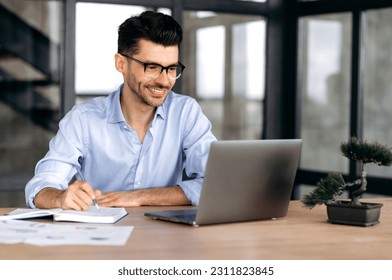 Online education. Intelligent successful caucasian man with glasses, sitting at the workplace, using a laptop, takes notes, studying or working online, listening to a webinar, gaining knowledge, smile - Shutterstock ID 2311823845