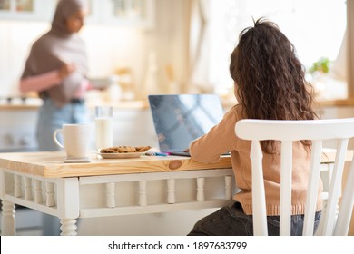 Online Education. Little Girl Study With Laptop In Kitchen While Her Muslim Mom Cooking Food, Small Female Child Sitting At Table With Computer, Having Distance Lesson With Teacher, Rear View