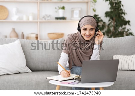 Online Education For Muslim Women. Happy Arabic Girl In Headscarf And Headset Studying With Laptop At Home, Taking Notes While Watching Webinar