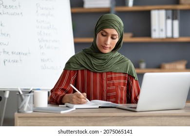 Online Education. Portrait of muslim woman in headscarf sitting at table writing in notebook with pen, using pc computer having video training during quarantine due to covid-19 coronavirus pandemic
