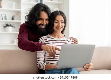 Online education, remote work and shopping. Happy indian couple looking at laptop and choosing purchase or watching lesson, sitting on couch in living room at home, copy space