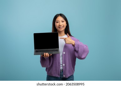 Online Educational Courses. Cheerful Asian Female Student Pointing At Laptop With Empty Blank Screen And Smiling Over Blue Studio Background. Excited Teen Lady With Pc Computer, Mockup For Design