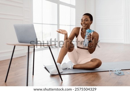 Online Exercise Session Marathon, Remote Training. Happy millennial sporty lady sitting on mat and talking with coach, having videocall with teacher, taking break from virtual workout drinking water
