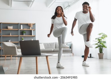 Online Family Workout. African American Husband And Wife Training Together In Living Room, Doing Twist Knees To Elbow Exercise. Happy Black Standing And Lifting Leg Up, Using Laptop, Watching Tutorial