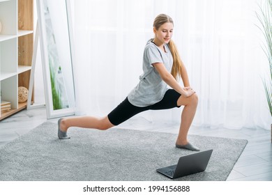 Online Fitness Training. Kid Home Sport. Workout Indoors. Active Lifestyle. Athletic Girl In Sportswear Doing Lunge Exercise Alone With Laptop In Light Room Interior.