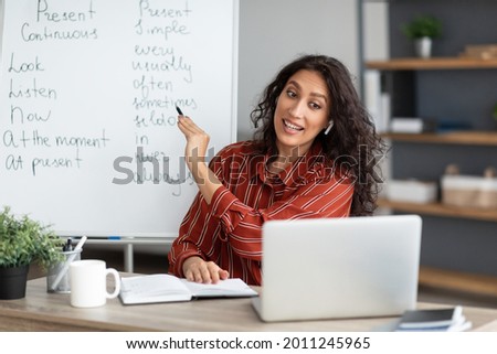Online Foreign Languages Tutoring. Joyful female teacher giving English class, pointing at blackboard with basic grammar rules. Experienced college professor explaining new material tenses to students