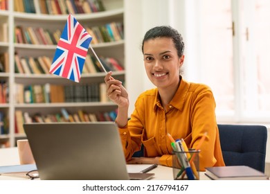 Online Foreign Languages Tutoring. Positive Female Teacher With Flag Of Great Britain Working With Laptop In Library Interior. Happy Lady Studying English Online For Abroad Job