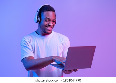 Online Gaming. Portraif Of Happy Black Man In Wireless Headphones Using Laptop Computer, Playing Games In Internet While Standing In Neon Lighting Over Purple Studio Background, Copy Space