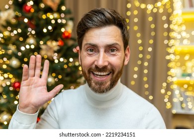 Online Greetings Video Call, Christmas Man Looks Into Web Camera And Smiles And Waves While Sitting On Sofa At Home Celebrating New Year Holidays.