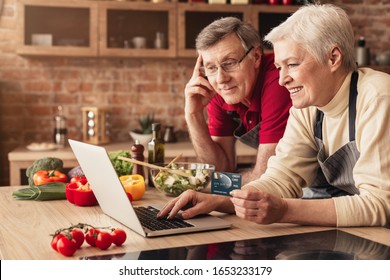 Online Grocery Concept. Happy Senior Couple Using Laptop And Credit Card In Kitchen, Ordering Food Delivery.