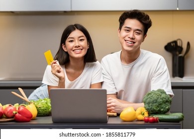 Online Grocery Shopping. Asian Couple At Laptop Showing Credit Card Posing In Modern Kitchen At Home, Smiling To Camera. Chinese Family Ordering Food Using Delivery Service On Weekend