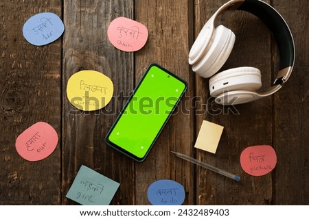 Online Hindi courses mobile app mockup. Cards with translated Hindi words, headphones and smartphone with green screen on wooden table.