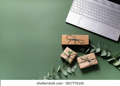 Online holiday shopping concept. Lap top, gift boxes and eucalyptus twigs on deep green background.