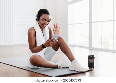 Online Home Training. Sporty black woman waving to cell phone webcam, sitting on the floor on yoga mat wearing wireless headset. Lady exercising in living room, making video call with personal trainer