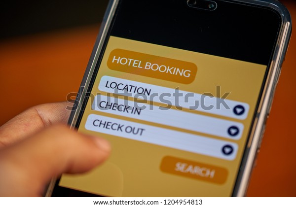 Online hotel booking concept. Male hands using\
smartphone for hotel reservation on mobile travel app with wood\
table background. Front view & close up. All screen graphics\
are made up with own\
design