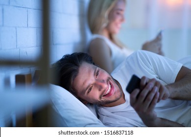 Online Infidelity. Husband Chatting On Smartphone Lying In Bed With Wife At Night. Low Light, Selective Focus