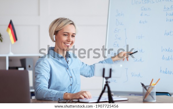 Online languages school. Millennial female tutor\
giving German lesson on internet, using smartphone, pointing at\
blackboard, explaining grammar to remote students. Education during\
covid-19 epidemic