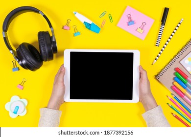 Online learning, back to school, remote education at home. Kid hands holding digital tablet computer. Headphones and school supplies on yellow background. Top view