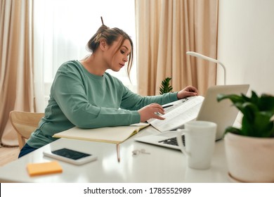 Apprentissage en ligne. Jeune belle étudiante en vêtements décontractés assise au bureau et lisant un manuel, utilisant son ordinateur portable, prenant des notes pendant ses études à la maison. Étude en ligne. Éducation à distance