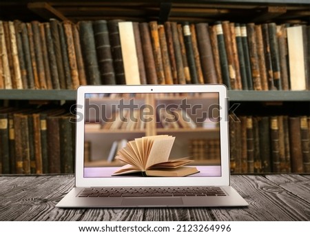 Online library. Modern laptop on wooden table and shelves with books indoors