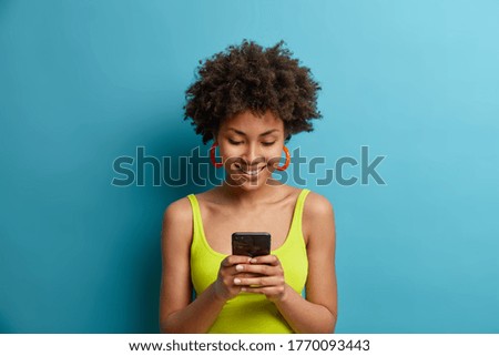 Online lifestyle and technology concept. Smiling charming woman looks at smartphone with beaming smile, stays in touch with boyfriend abroad, gazes satisfied in display, wears summer clothes