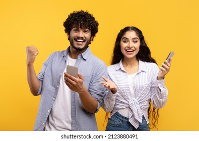 Online Lottery. Excited Young Indian Guy And Lady Rejoicing Success And Holding Smartphones, Exclaiming With Triumph While Standing On Yellow Background