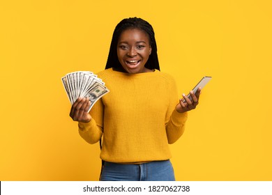 Online Lottery. Joyful Black Lady Holding Smartphone And Dollar Cash, Celebrating Success With Bunch Of Money, Lucky Female Received Big Prize, Standing Isolated On Yellow Background With Copy Space