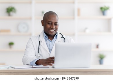 Online Medical Consultation. Highly Qualified Black Doctor Consultating Patients Distantly From His Office During Quarantine, Using Laptop Computer, Typing Diagnosis And Sending Emails