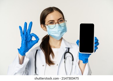 Online Medical Help Concept. Enthusiastic Young Woman Doctor In Face Mask, Showing Okay Sign And Mobile Phone App, Smartphone Screen Interface, White Background