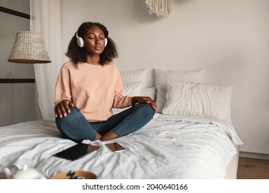 Online Meditation. Peaceful Black Woman Meditating With Eyes Closed Wearing Headphones Sitting In Lotus Position, Listening To Yoga Instructor Via Digital Tablet Relaxing In Bedroom At Home