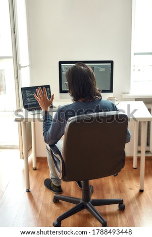 Online meeting with colleagues. Back view of male web developer looking at webcam and waving while sitting at his workplace and working from home, writing code. Freelance. Stay home, self isolation