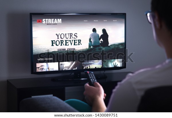 Online movie stream service in smart tv. Streaming\
series with on demand video (VOD) service in television. Man\
choosing film to watch with remote. Person sitting on couch at home\
late at night.