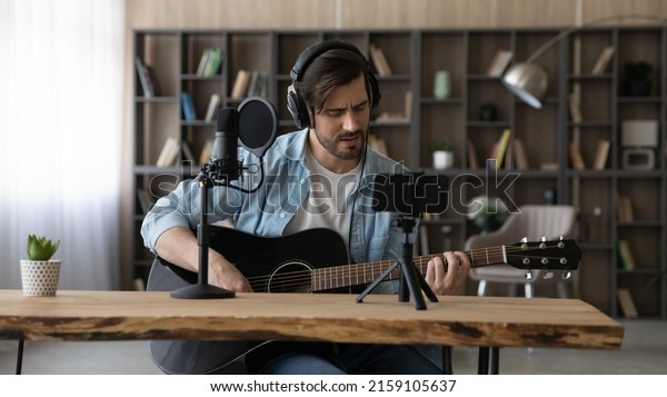 Online music lesson. Bearded millennial male\
teacher wear headphones sit by desk with digital microphone on\
holder give web video consultation using phone webcam teach student\
to play acoustic guitar
